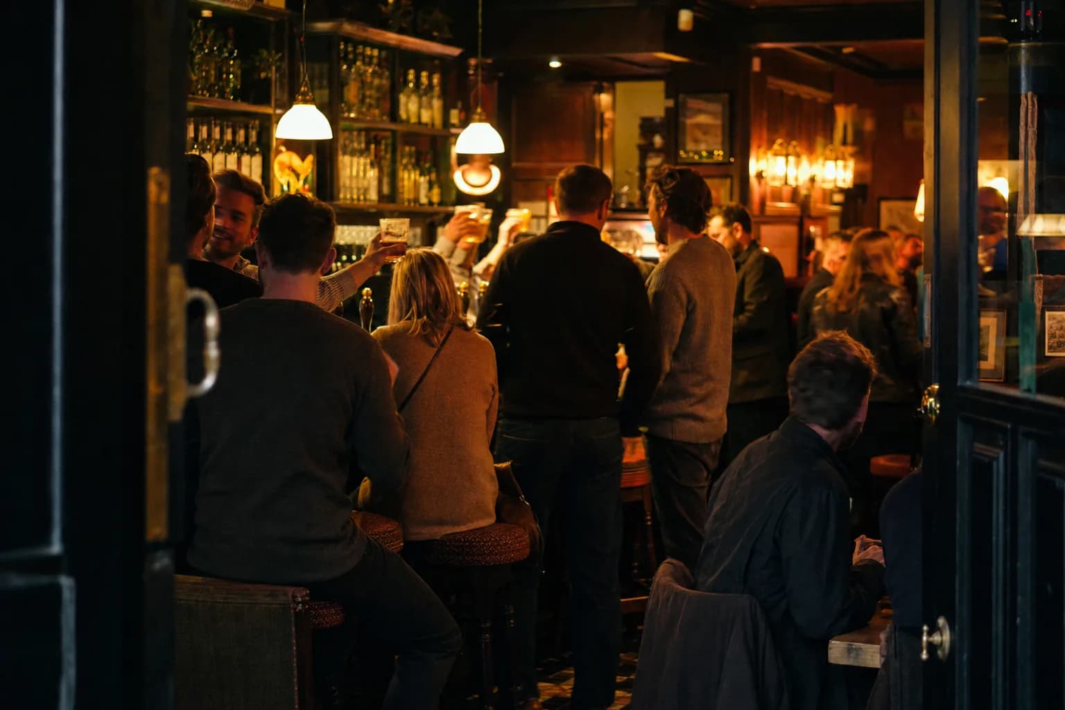 A busy pub bar at night, seen from the doorway, glasses raised (AI example photo)
